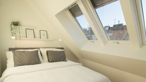 A bedroom with a sloped ceiling, a bed with white bedding, two pillows, a bedside table with a lamp, and a view of buildings through a skylight. - Stylish & Modern Apartment (Vienna)