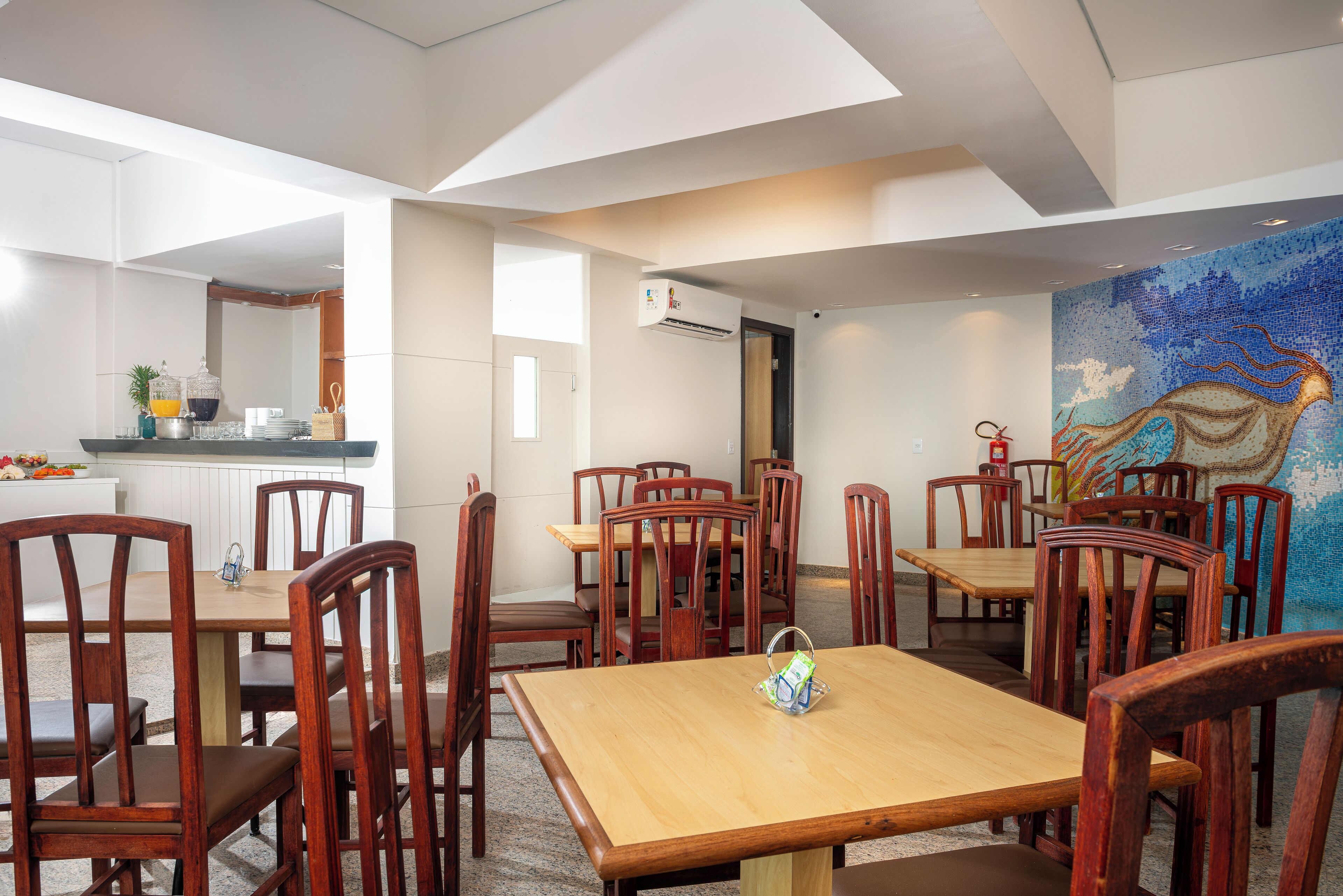 A dining area with wooden tables and chairs, a kitchen in the background, and a colorful mural on the wall.