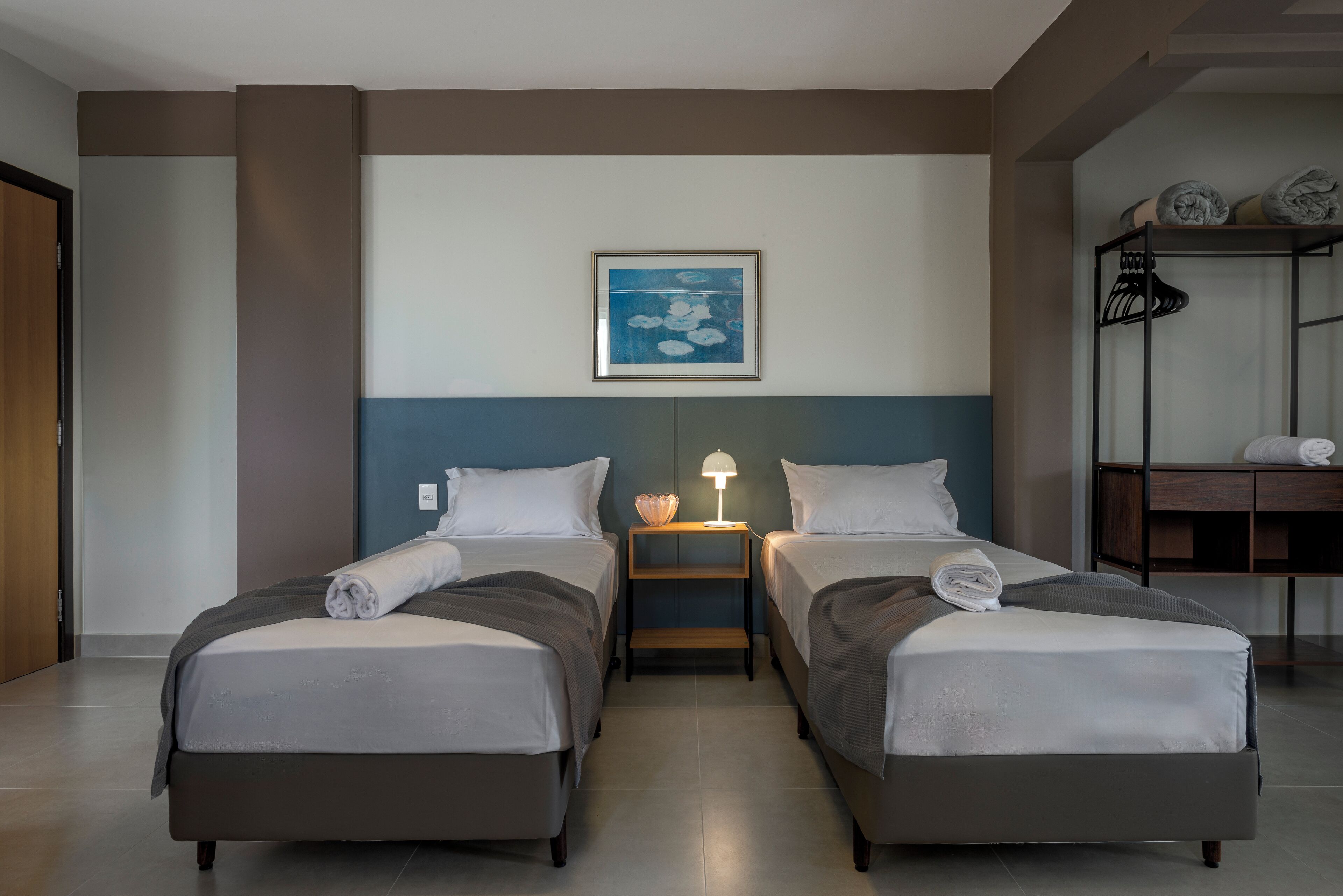 Habitación de hotel con dos camas individuales, una mesita de noche con lámpara, un cuadro en la pared y un estante de madera con toallas dobladas.