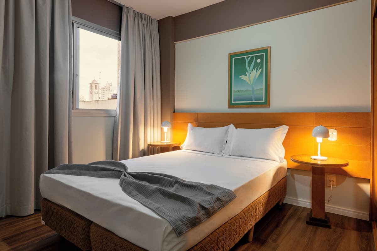 A hotel room with a large bed, two bedside lamps, a window with curtains, and a painting on the wall.