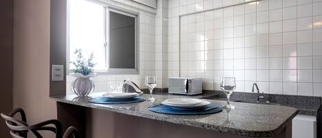 A kitchen with white tiled walls, a granite countertop, a microwave, a sink, and a window with blinds.