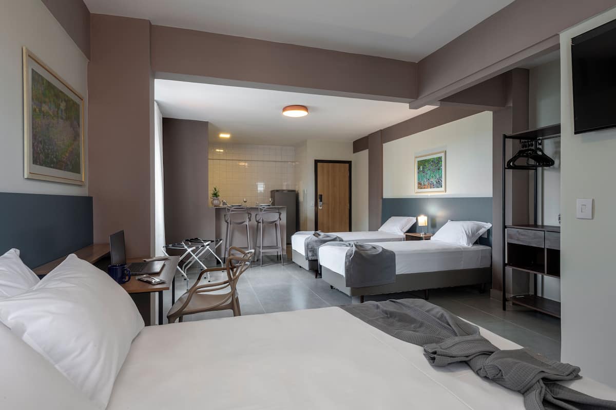 A hotel room with a large bed, a desk with a laptop, a chair, and a small kitchenette in the background.