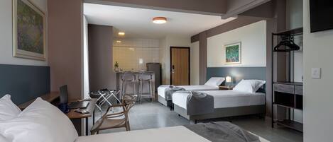 A hotel room with a large bed, a desk with a laptop, a chair, and a small kitchenette in the background.