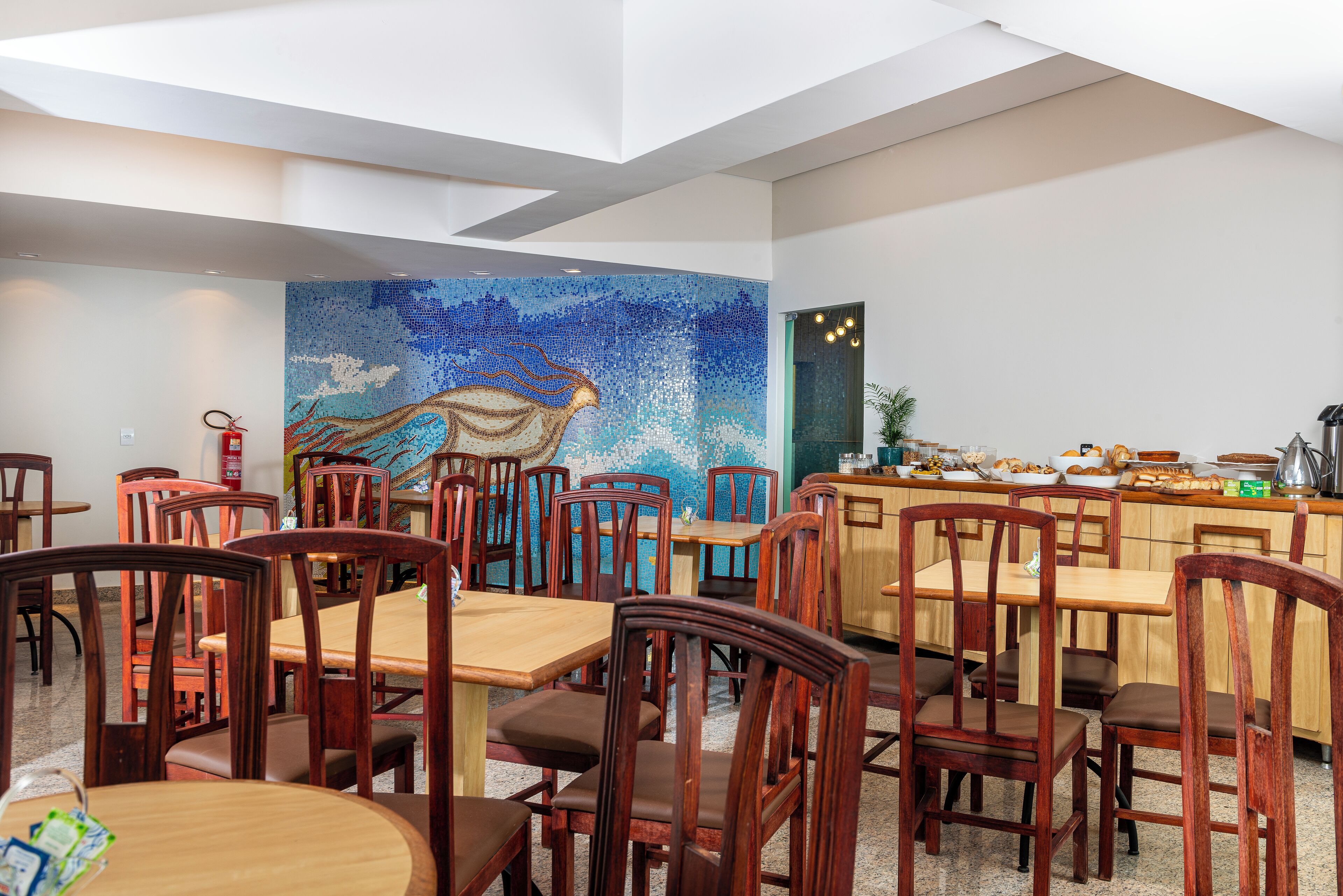En restaurant med træborde og stole, en mosaikvæg og et køkkenområde med forskellige madvarer.