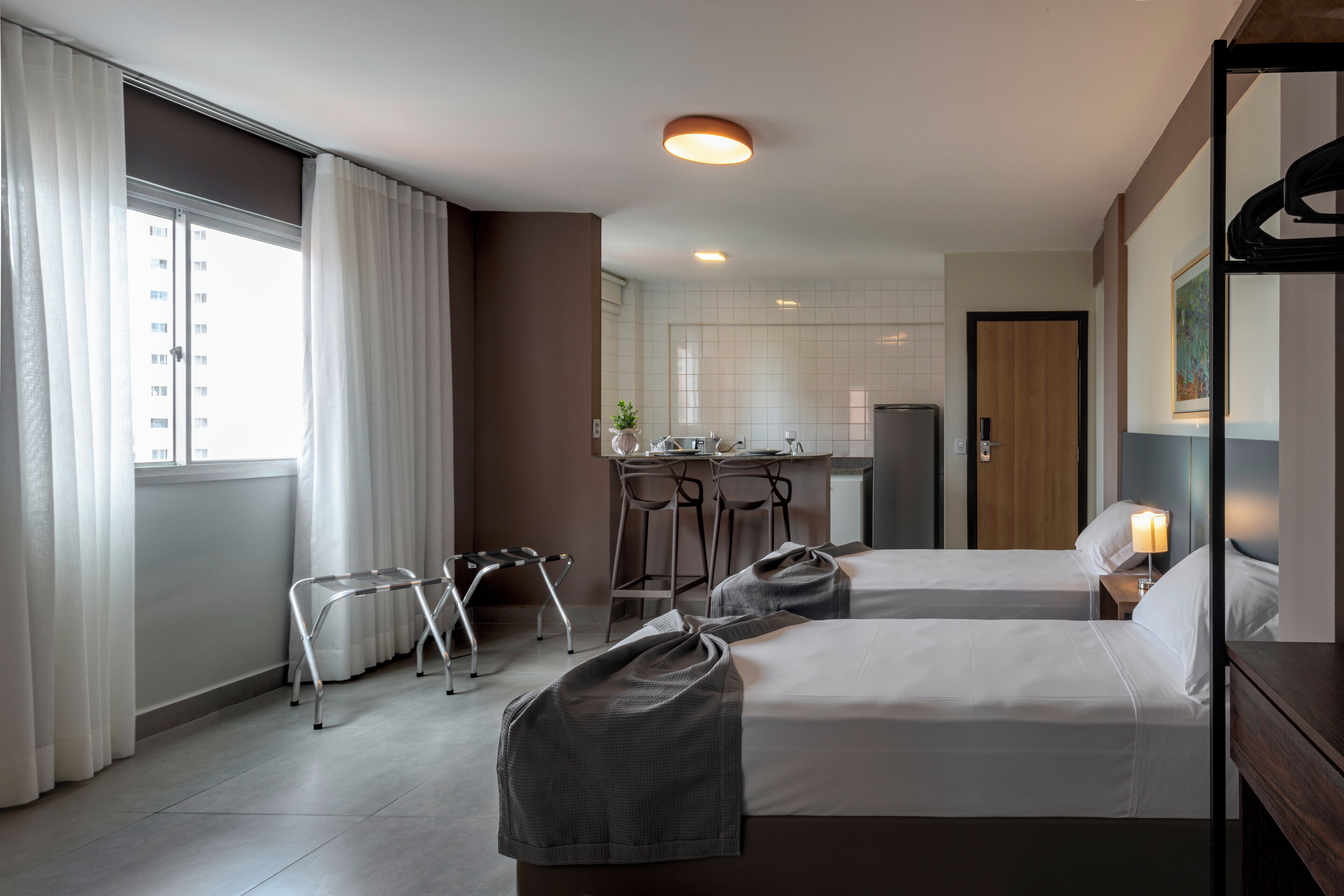 A hotel room with two beds, a kitchenette, a dining area, and a view of a building outside.