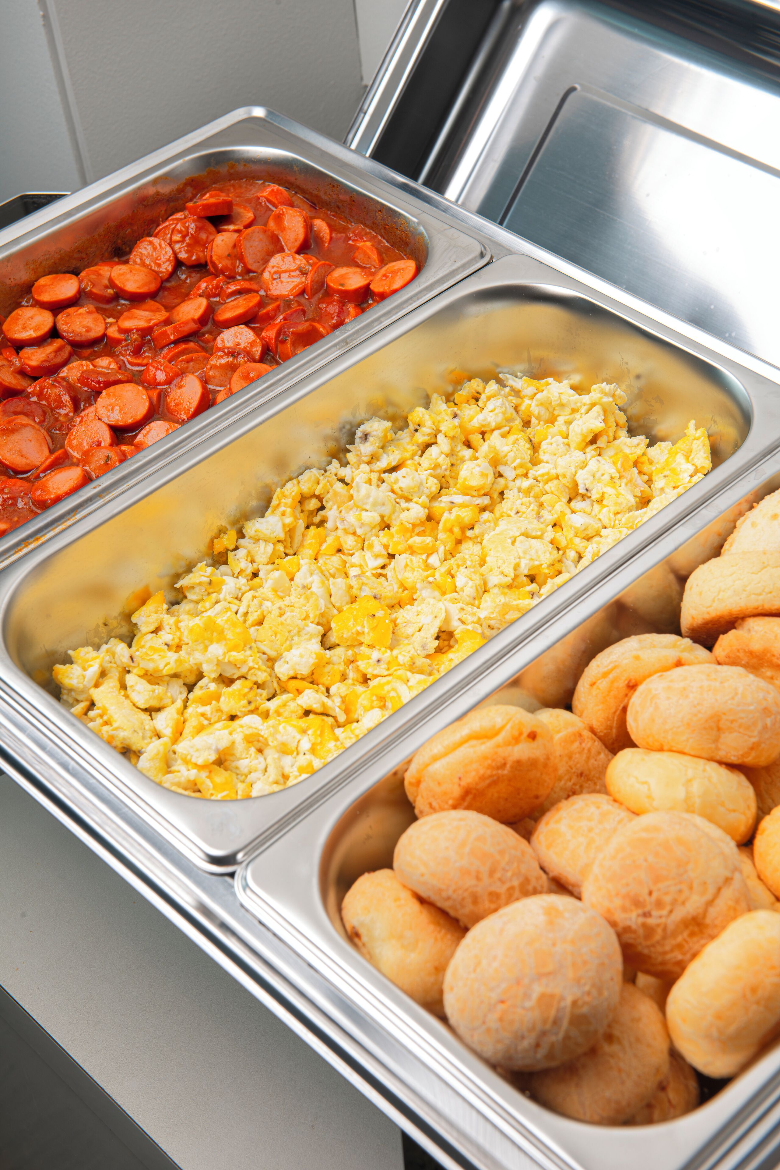 Un bufé con tres fuentes que contienen huevos revueltos, rebanadas de salchicha y bolitas de queso.