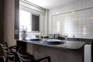 A kitchen with white tiled walls, a granite countertop, a microwave, a sink, and a window with blinds. - Lym Flat Hotel (Uberlandia)