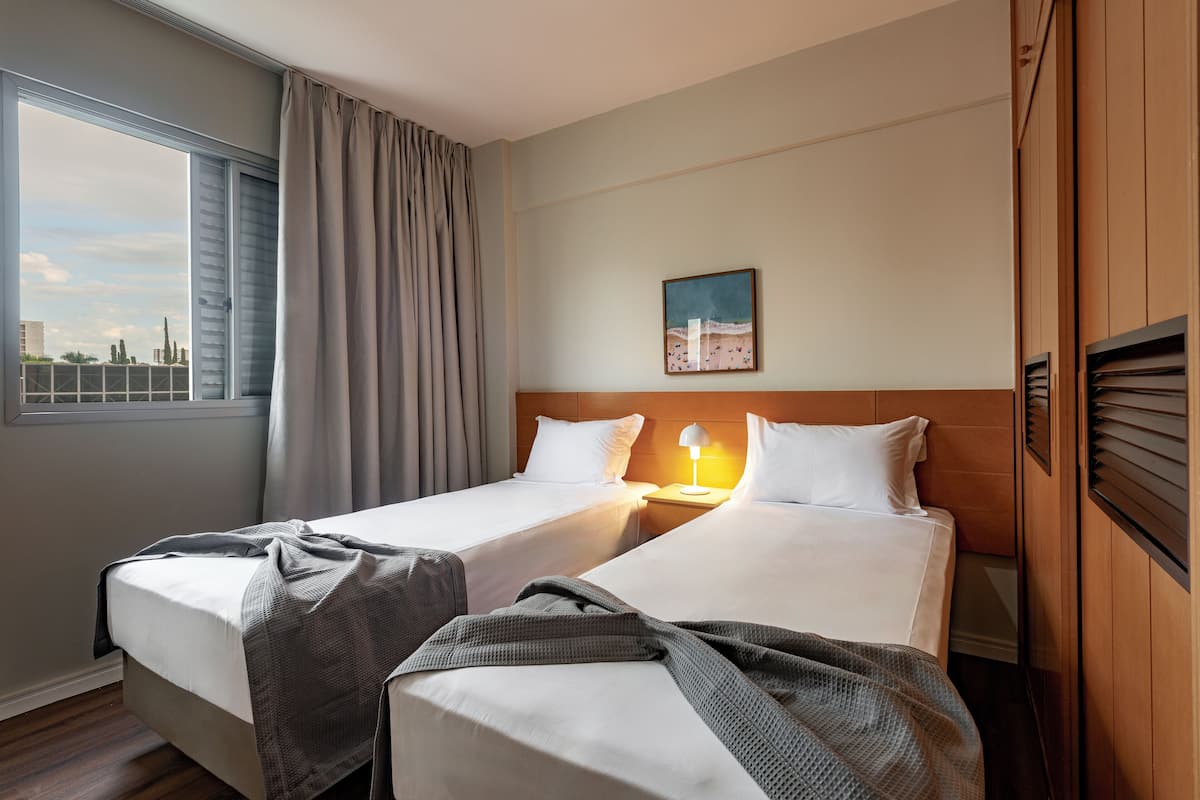 A hotel room with two beds, a window with curtains, a painting on the wall, and a wooden headboard.