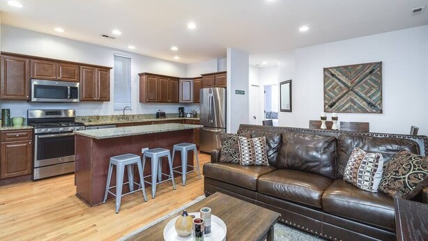 Una cocina moderna con electrodomésticos de acero inoxidable, una isla central con taburetes y un sofá de cuero marrón en una sala de estar con un gran elemento decorativo en la pared.