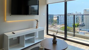 Een moderne woonkamer met een flatscreen televisie aan de muur, een witte tv-kast, een ronde houten tafel met een vaas, en een groot raam met uitzicht over de stad.