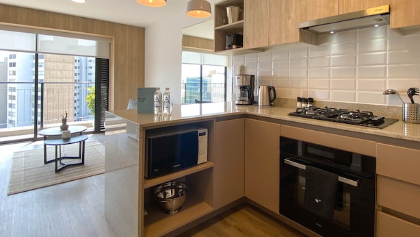 Een moderne keuken met houten kastjes, een ingebouwde magnetron, een fornuis en een koffiezetapparaat. Er is een klein zithoekje met een tafel en stoelen, en er is uitzicht op de stad door grote ramen.