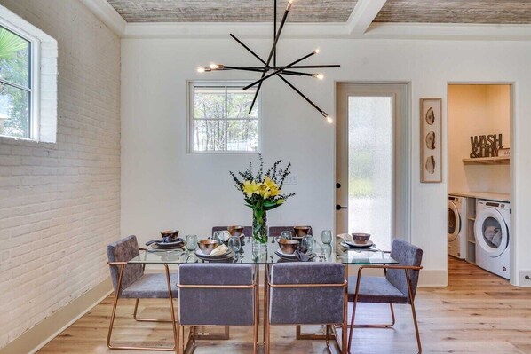 Un comedor con mesa de vidrio, cuatro sillas, una lámpara moderna y vista a un cuarto de lavado.