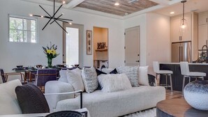A modern living room with a grey sofa, a dining area with a table and chairs, and a kitchen with a refrigerator and pendant lights.