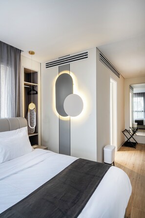 Una habitación de hotel moderna con una cama grande, un armario, un espejo con iluminación integrada, una mesita y una ventana con cortinas.