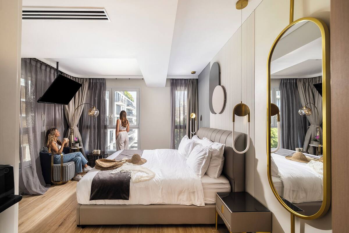 A modern hotel room with a large bed, a bedside table, a mirror, and a view of the outside.