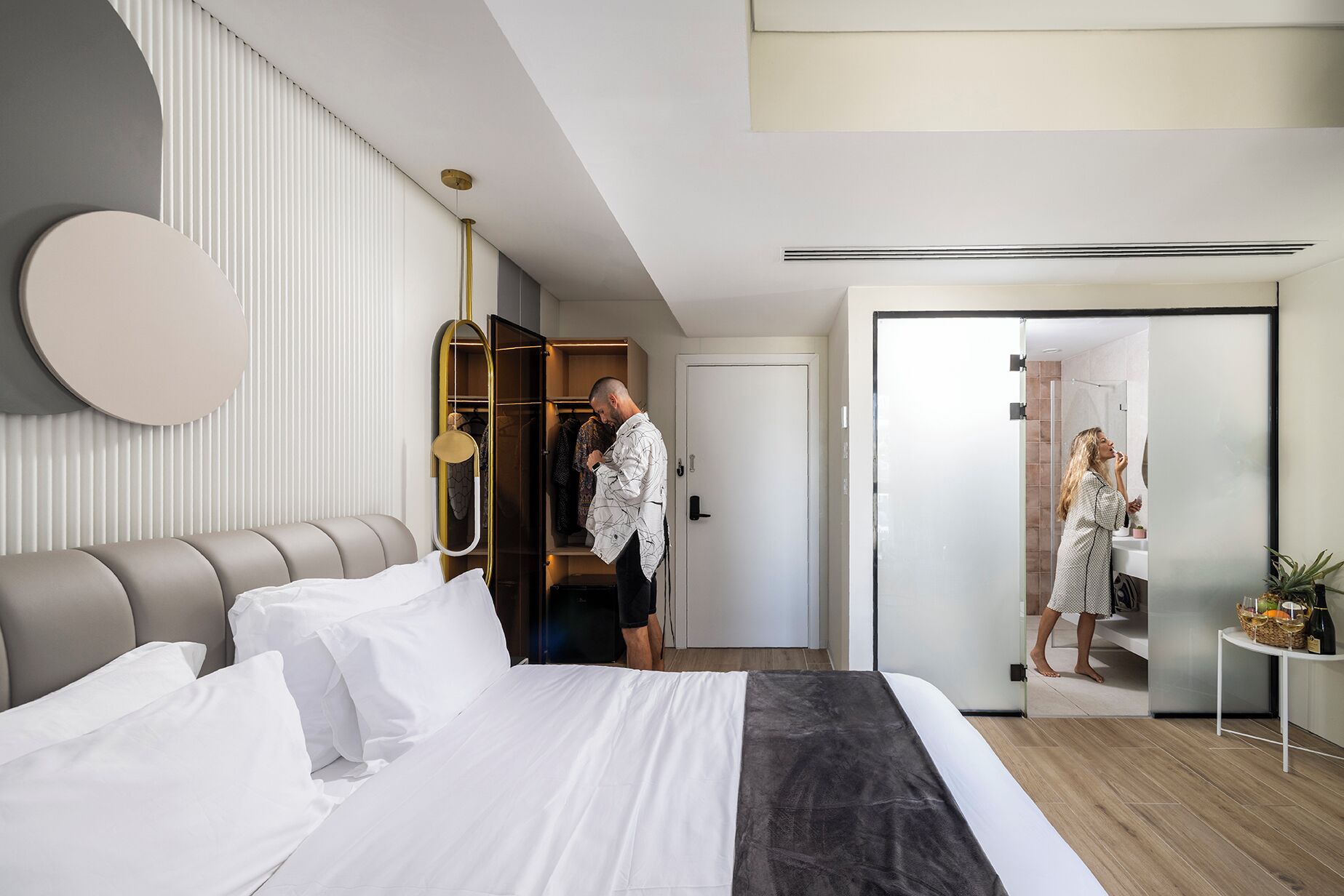Una habitación de hotel moderna con una cama, un vestidor, un baño con espejo y una mesita con una planta.
