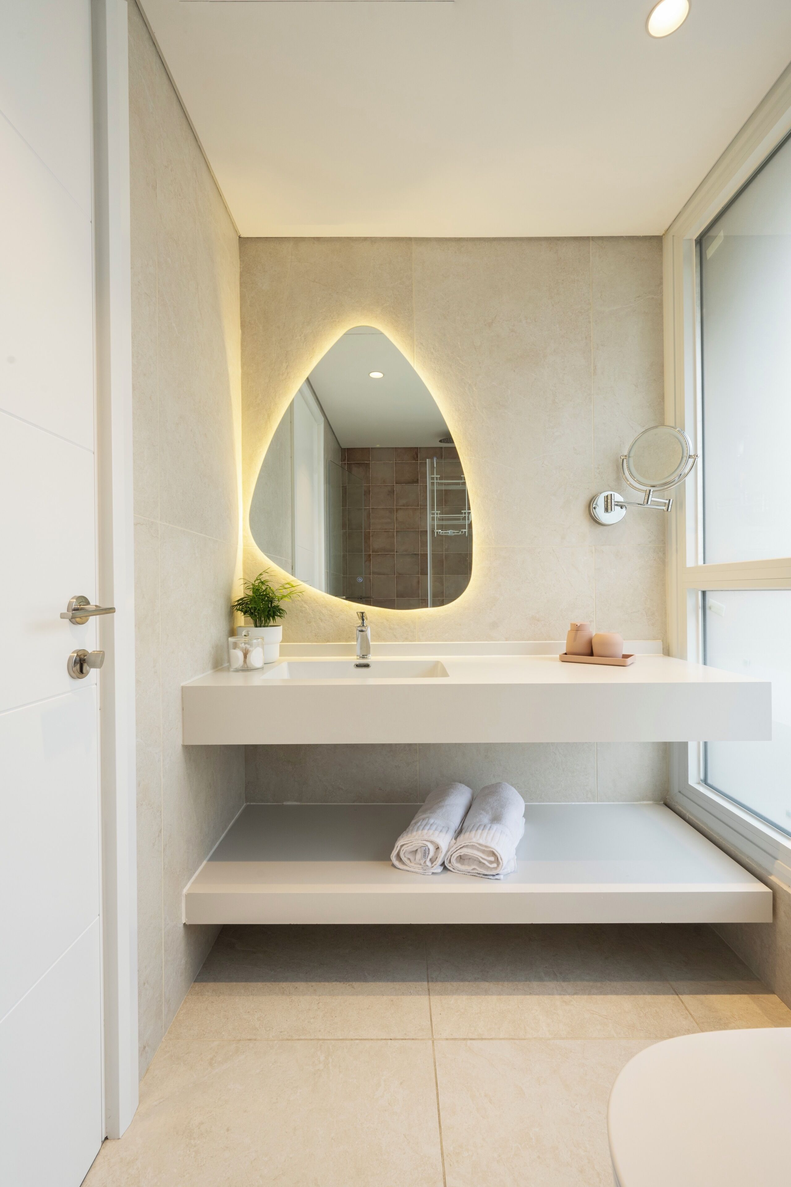 Un bagno moderno con un lavello integrato, uno specchio dalla forma particolare, una piccola pianta e un portasacchi.