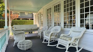 Une véranda meublée de mobilier en osier, comprenant un canapé et des fauteuils à bascule, ainsi qu’une cage à oiseaux blanche.