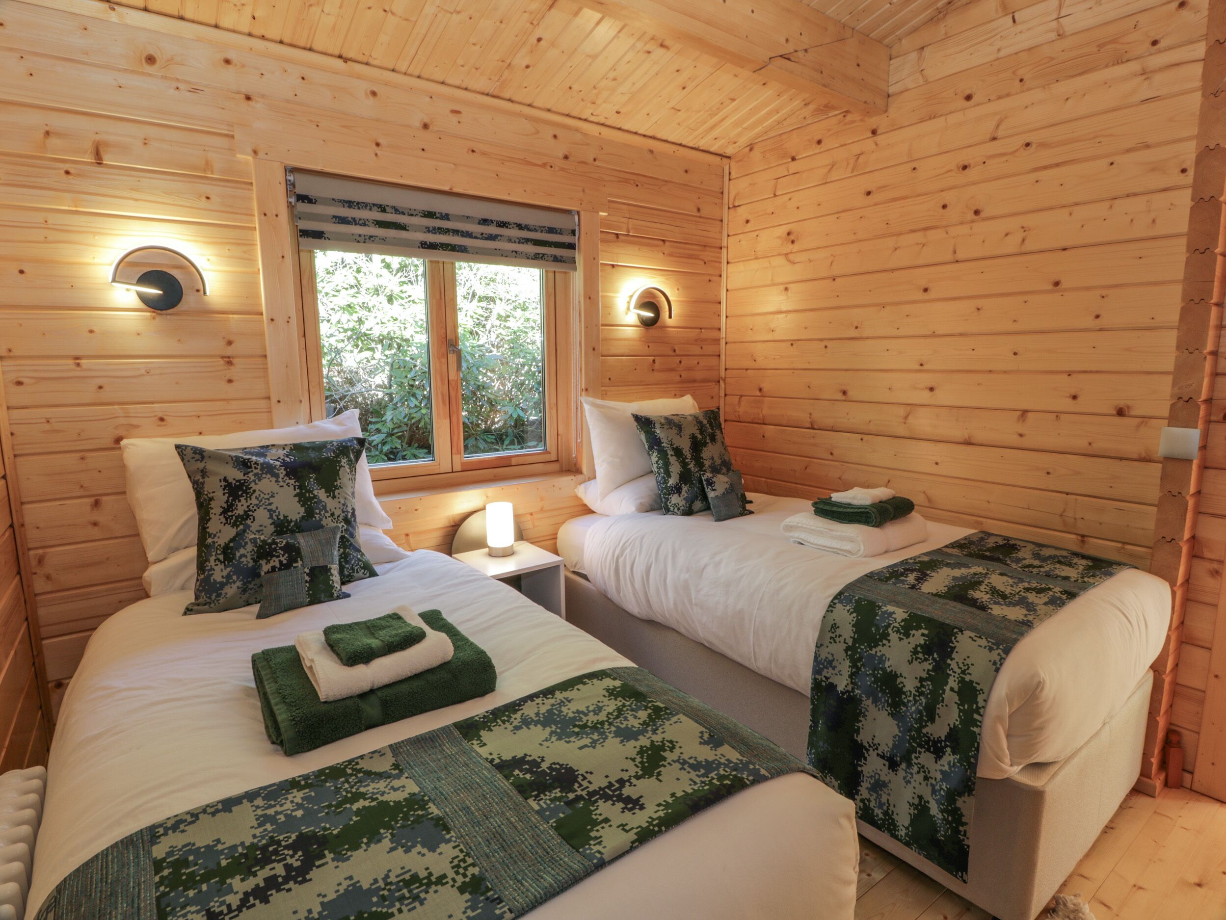 Habitación de cabaña de madera con dos camas, cada una con una manta verde de camuflaje, una mesita de noche y un ventanal con vistas a la vegetación.
