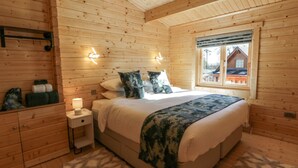 A wooden cabin bedroom with a bed, bedside tables, and a window with a view of another cabin. - Little Lodge (Newton Stewart)