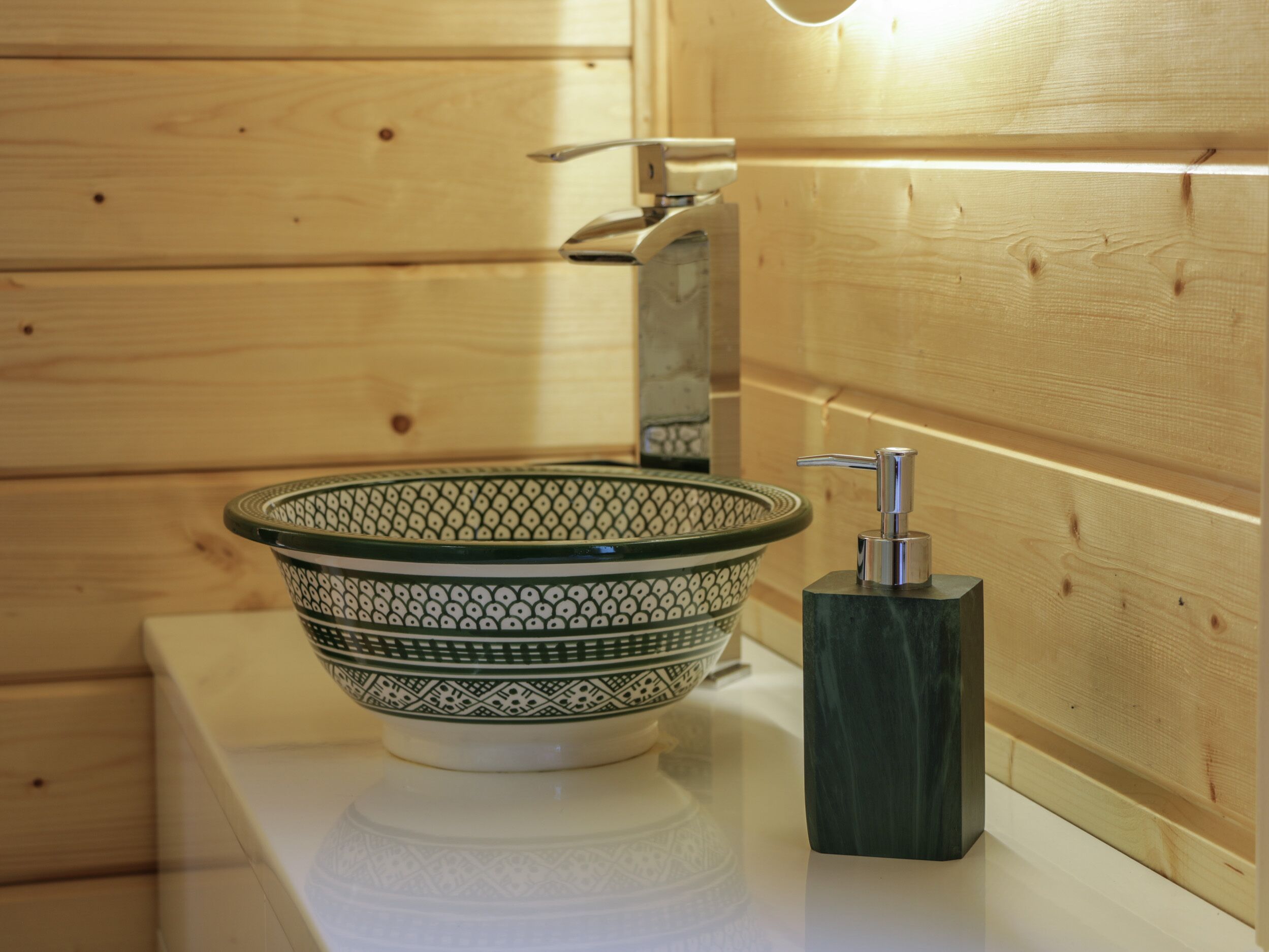 Une salle de bain avec un mur en bois, un comptoir blanc, un lavabo décoré de motifs verts et blancs, et un distributeur de savon noir.