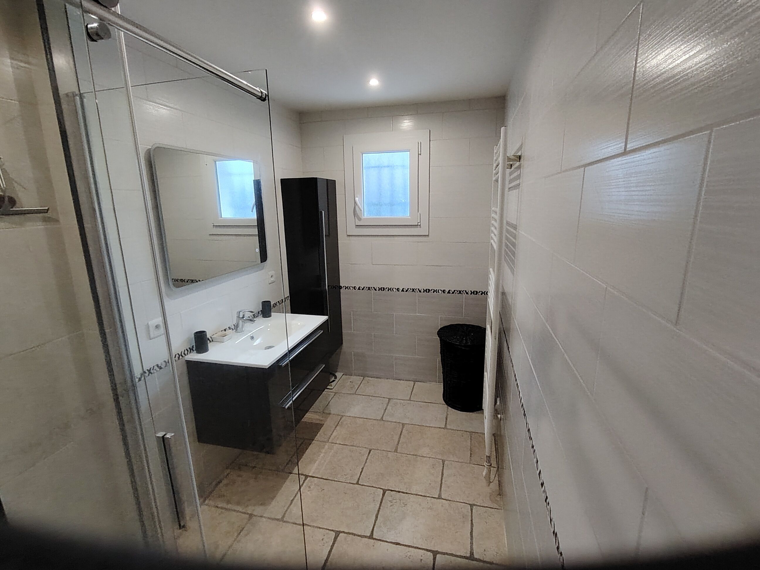 Een badkamer met een glazen douchecabine, een witte wastafel, een zwart kastje en een zwarte prullenbak.
