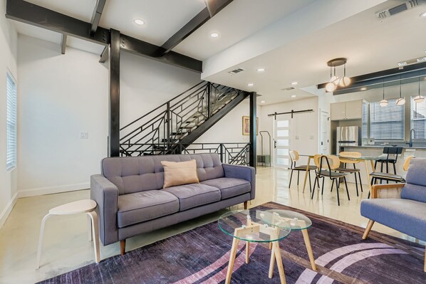 Una sala de estar moderna con un sofá gris, una mesa de centro de vidrio y una escalera que sube a un nivel superior.