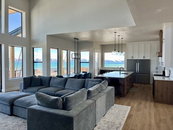 Een moderne woonkamer met een grijze hoekbank, een keuken met witte kastjes, en grote ramen met uitzicht op zee en bergen.