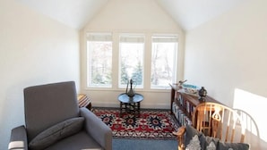 A cozy room with a sloped ceiling, a gray armchair, a patterned rug, a small round table with a vase, and a bookshelf with books and decorative items. - Peaceful Retreat (Minneapolis)