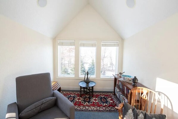 A cozy room with a sloped ceiling, a gray armchair, a patterned rug, a small round table with a vase, and a bookshelf with books and decorative items.