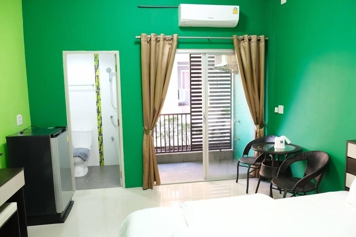 A hotel room with a green wall, a bed, a desk, a chair, a table, a balcony, and a shower.