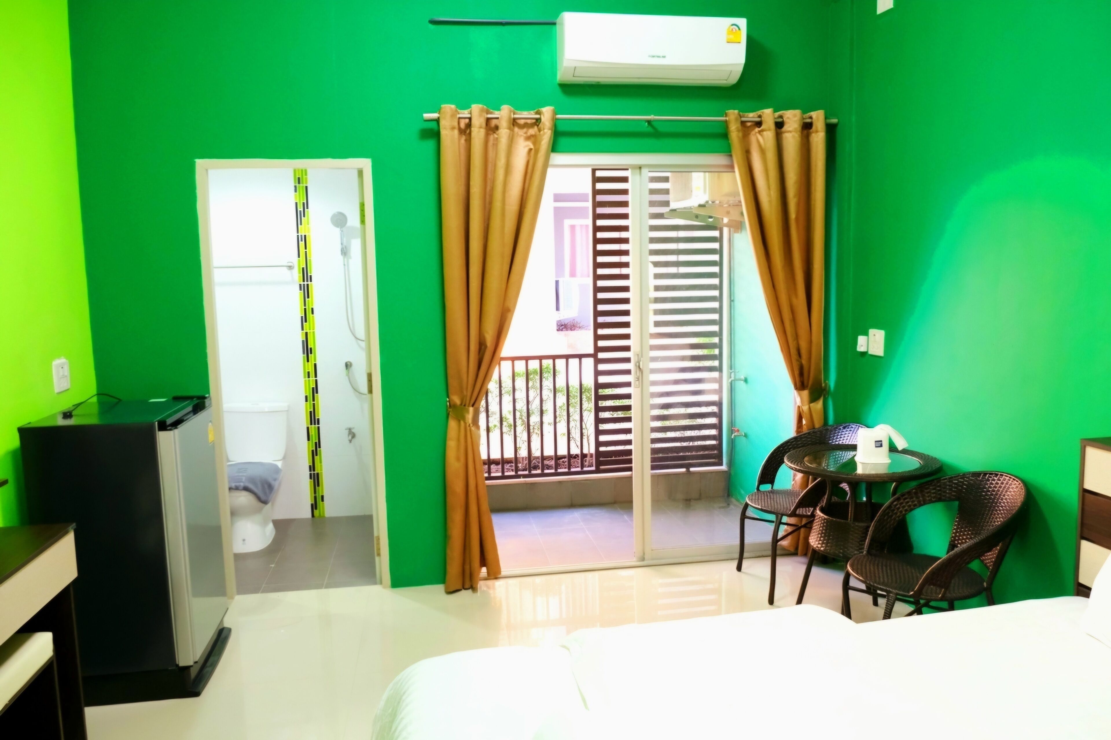 Habitación de hotel con pared verde, cama, escritorio con computadora, baño con ducha y balcón con barandilla.