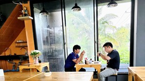 A restaurant with wooden tables and chairs, a kitchen area, and large windows with a view of greenery.