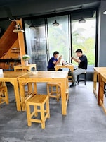 Un restaurante con mesas y sillas de madera, zona de cocina y grandes ventanales con vistas a zonas verdes.
