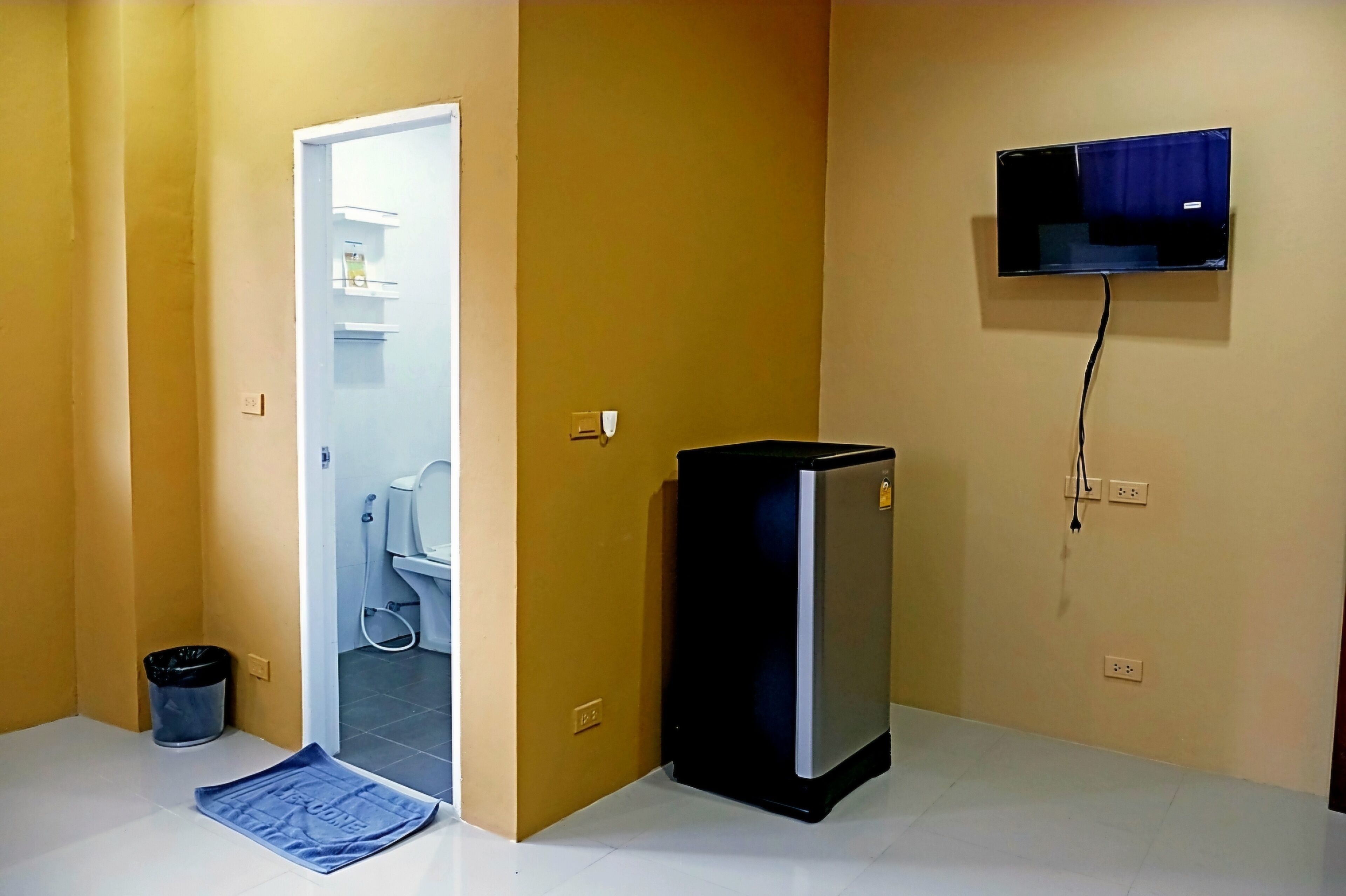 Una habitación pequeña con una puerta que da a un baño, un televisor montado en la pared y un refrigerador pequeño.