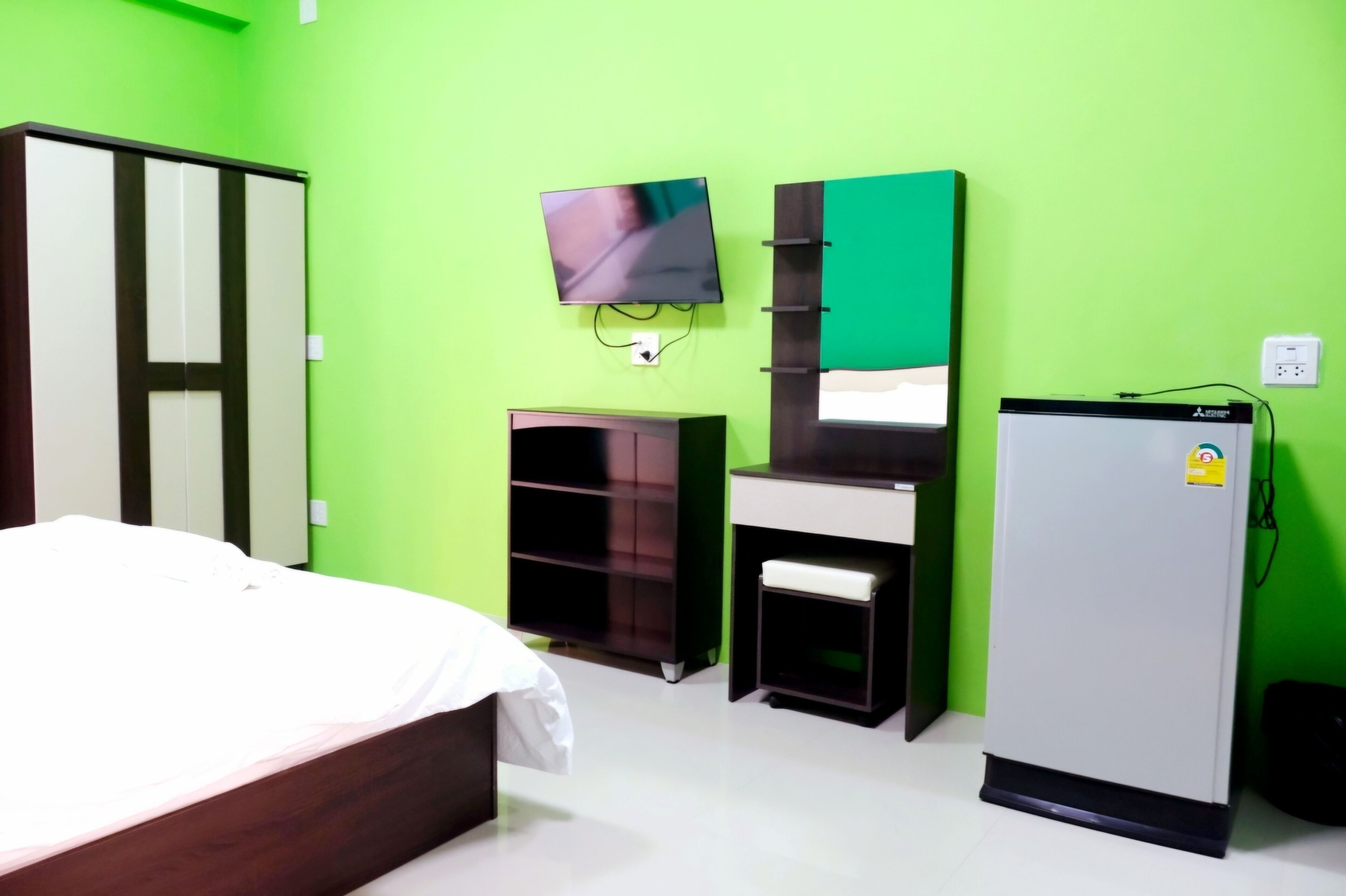 A room with a bed, a TV mounted on the wall, a wardrobe, a desk with a chair, and a refrigerator.