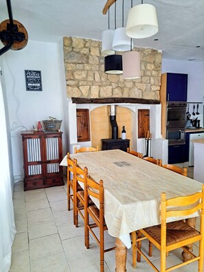A dining area with a long table, wooden chairs, a stone wall, and a kitchen in the background. - Former forge, leisure area with pool (Marigny)