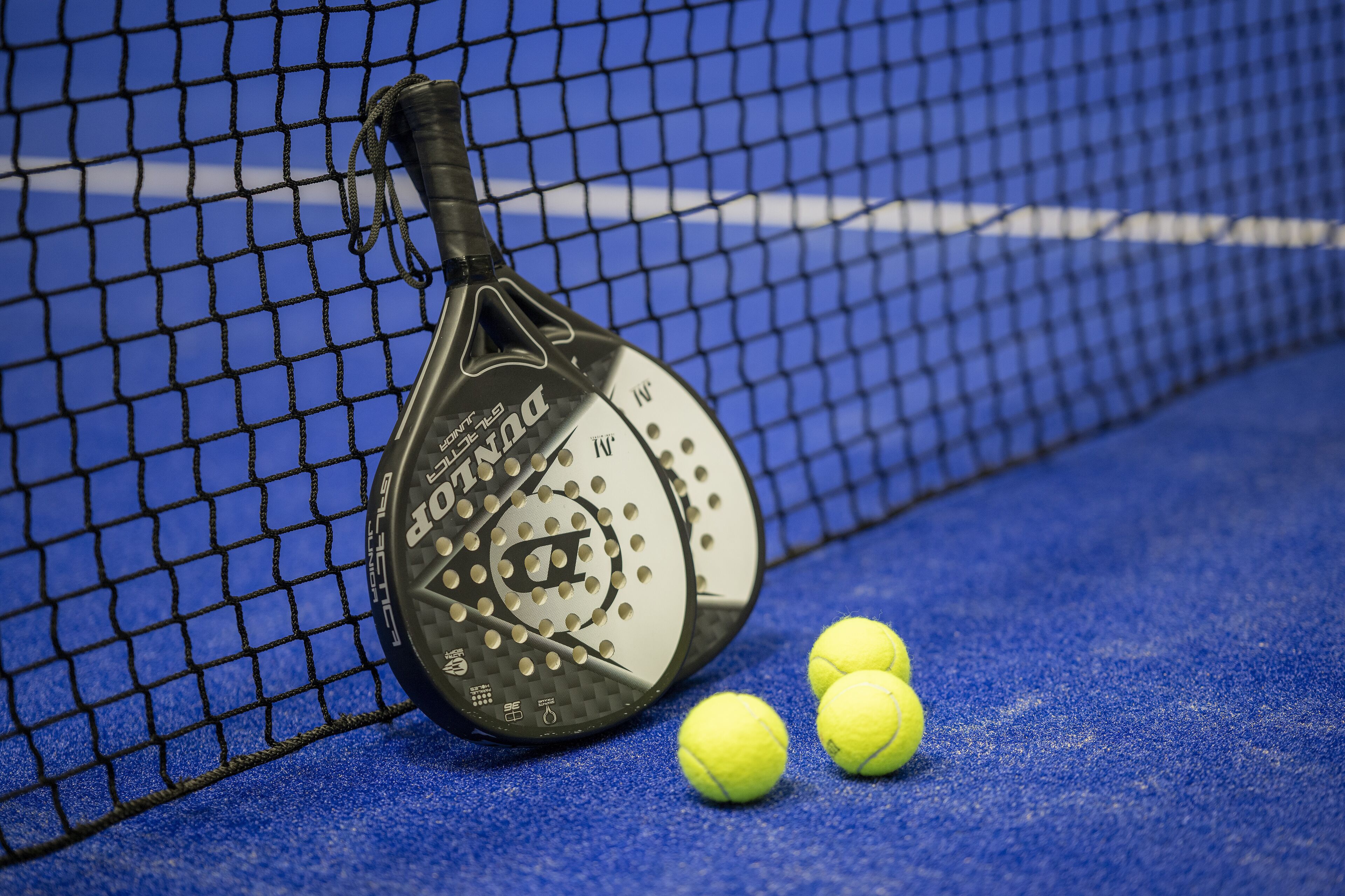 Une raquette de tennis et trois balles de tennis sur une surface bleue, avec un filet en arrière-plan.