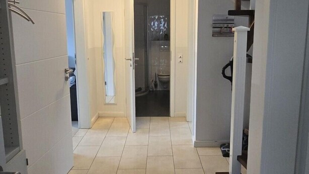 A modern hallway with a staircase leading to a second floor. The floor is tiled, and there are white walls with a door at the end. - Strandkrabbe: Your vacation by the sea! (Borkum)