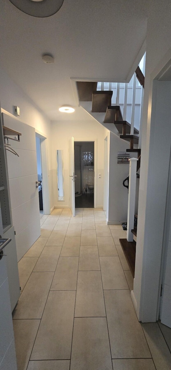 Ein moderner Flur mit einer Treppe, die zu einer zweiten Etage führt. Der Boden ist mit Fliesen belegt und die Wände sind weiß, am Ende befindet sich eine Tür.