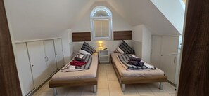 A small, narrow room with two single beds, a nightstand, and a window with blinds. - Strandkrabbe: Your vacation by the sea! (Borkum)