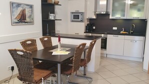 A modern kitchen with white cabinets, a black countertop, and a dining area with wicker chairs and a table. - Ferienwohnung Borkumgarten (Borkum)