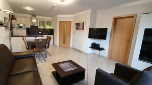 A modern living room with a sofa, armchair, and coffee table. There is a dining area with wicker chairs and a small kitchen with a microwave and a TV. - Ferienwohnung Borkumgarten (Borkum)