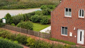 A red brick house with a fenced yard, a car parked in the driveway, and a grassy area with bushes. - Ferienwohnung Borkumgarten (Borkum)