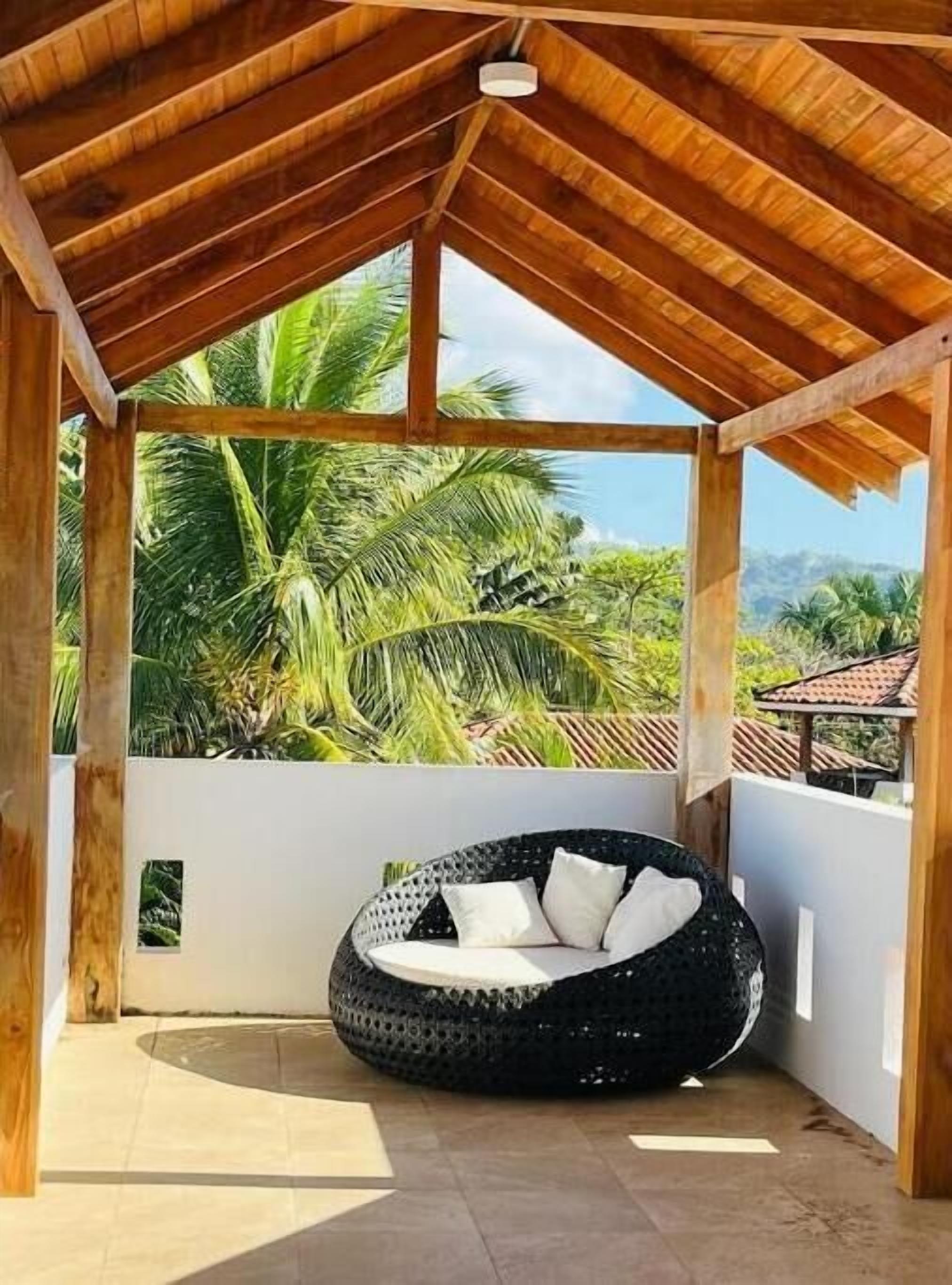 Un espace extérieur aménagé avec des sièges, protégé par un auvent, comprenant une chaise en osier noir et des coussins, le tout entouré de palmiers et offrant une vue dégagée sur le ciel.
