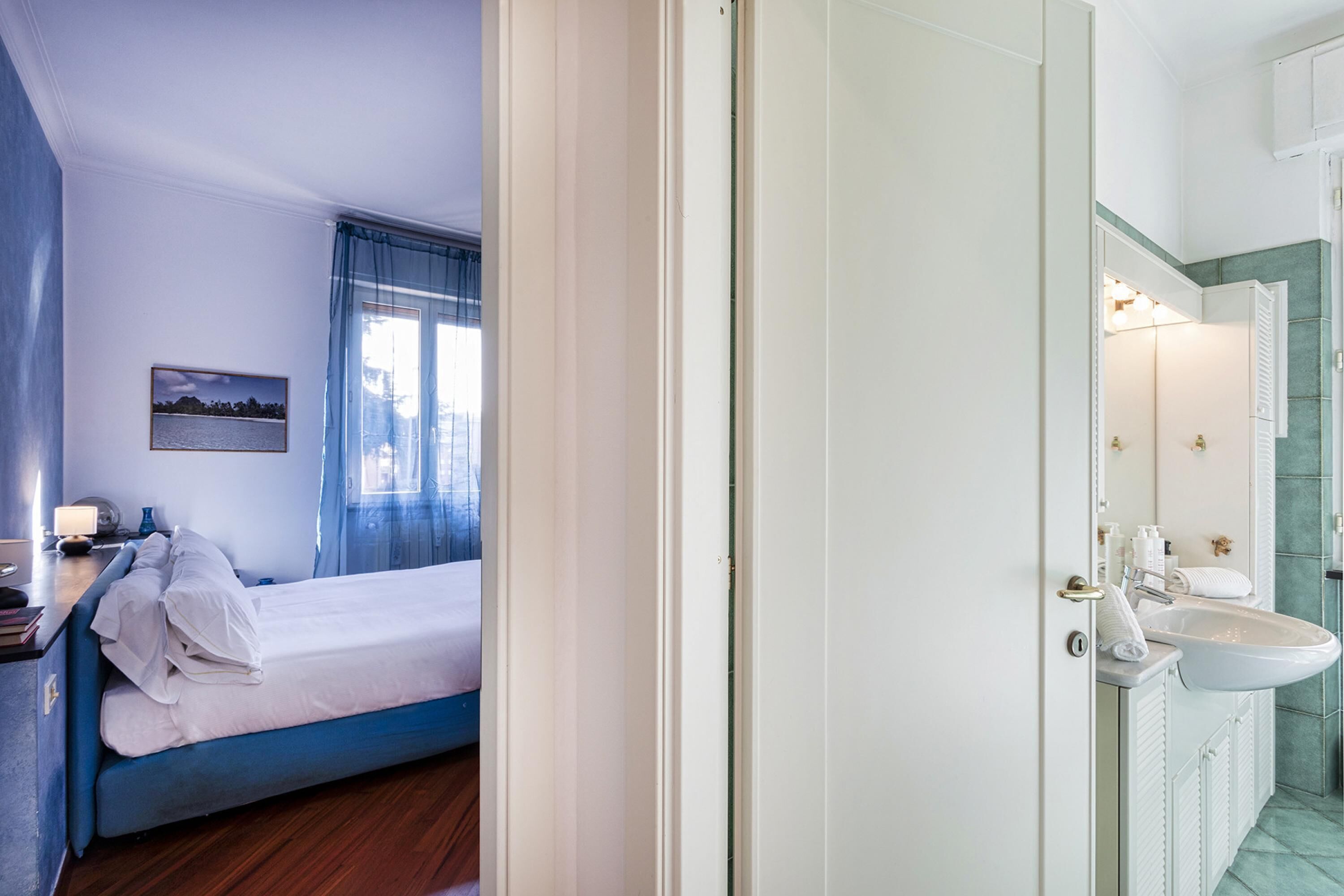 Quarto de hotel com cama, mesa de cabeceira com abajur, janela com cortinas e banheiro com pia e espelho.