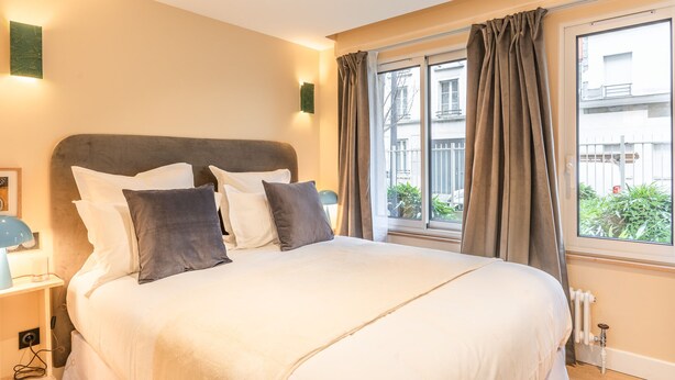 Un lit bien fait, avec une literie blanche et grise, une table de chevet avec une lampe et une fenêtre avec des rideaux.