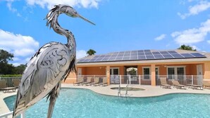 Ein Pool mit einer großen Vogel-Skulptur, ein Gebäude mit Solaranlagen und ein eingezäunter Bereich mit Liegestühlen.