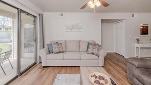 Ein modernes Wohnzimmer mit einem beigen Sofa, einem Deckenventilator aus Holz und einer Glastür, die auf eine Terrasse führt.