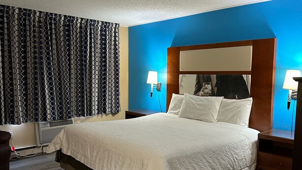Una habitación de hotel con una cama grande, una mesita de noche con lámpara, una silla y aire acondicionado instalado en la pared.